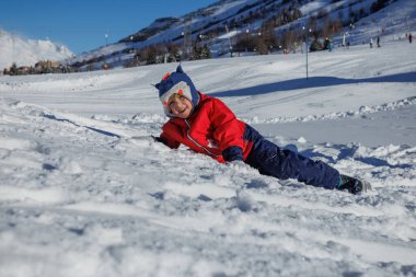 Canlı kırmızı ceketli bir çocuk açık bir kış gününde karlı bir yamaçta yatmaktan hoşlanır.