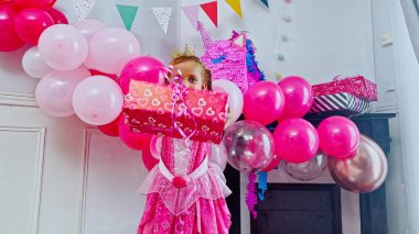 Pembe prenses kostümünün içinde gülümseyen kız, elinde kalp süslemeli bir hediye tutuyor, onu renkli balonlarla çevrili bir şekilde kameraya gösteriyor.