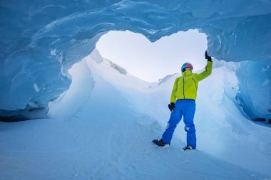 Canlı kıyafetli bir kayakçı görkemli bir buz mağarasında duruyor, gökyüzü yukarıdaki büyük kalp şeklindeki açıklıktan görülebiliyor.