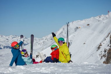 Parlak kayak kıyafetleri giymiş bir aile, arka planda karla kaplı dağlardan oluşan bir manzarayla karın üzerinde mutlu bir şekilde dinleniyor.