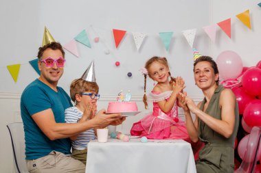 Bir aile, parti şapkaları ve neşeli aksanlarıyla süslü pembe bir pastayla doğum gününün tadını çıkarır.