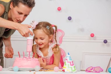 Bir kadın ve genç bir kız, renkli parti eşyalarıyla çevrili pembe bir doğum günü pastasını şekerle süslüyorlar.