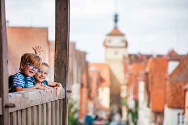 Rothenburg şehrinin ahşap bir balkonunda duran iki çocuk, arka planda eski bir kasaba ve saat kulesi ile samimi bir şekilde gülümsüyor.
