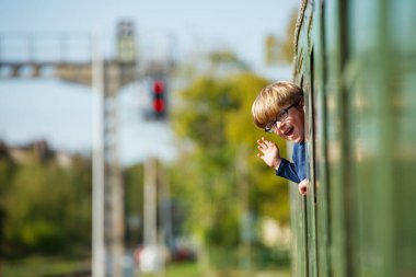 Camdan sarkan gözlüklü mutlu çocuk hareket halindeki yeşil vagondan el sallıyor.