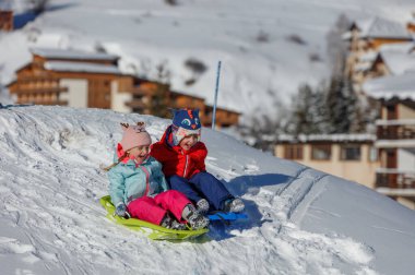 İki çocuk, kış manzarası ve arka plandaki evlerle çevrili karlı bir tepeden kayarak kayıyorlar.