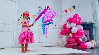 Pembe elbiseli genç bir kız gözleri bağlı ve renkli tek boynuzlu at pinatasına vurmaya hazırlanıyor. Dolu balonlarla dolu bir odada.