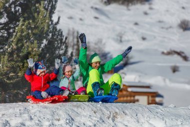 Kış kıyafetleri içinde üç çocuk çılgınca tezahürat yaparken, güneşli bir gökyüzünün altında kırmızı, yeşil ve mavi kızaklarda el kaldırırken.