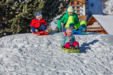 Parlak ceketli üç çocuk renkli kızaklarda karlı bir yamaçta oturuyor. Çam ağaçları çerçeveli ve uzaktaki tatil köyleri binaları.