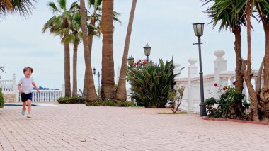 Küçük bir çocuk, palmiye ağaçları ve yemyeşil yapraklarla kaplı tuğla bir patika boyunca hevesle koşuyor, arka planda ışıklar var.