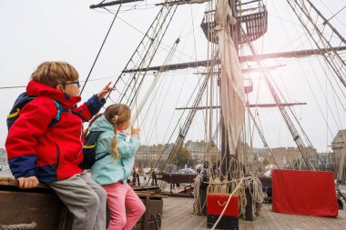 Kırmızı paltolu bir çocuk ve mavi ceketli bir kız büyük bir yelkenli gemisinin ahşap kutusunda, maceraya hazır bir şekilde oturuyorlar.