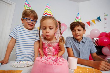 Parti şapkalı heyecanlı çocuklar doğum gününde yanan mumları söndürmek için pembe bir pasta etrafında toplanıyorlar.