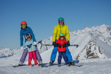 Renkli kıyafetler içinde, dört kişilik bir aile, anne, baba kız çocuk güneşli bir dağ yamacında kayak macerasından zevk alıyor.