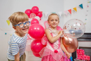 Gözlüklü ve çizgili tişörtlü genç bir çocuk doğum günü partisinde etrafında renkli balonlar olan pembe peri kostümlü bir kızın yanında gülüyor.