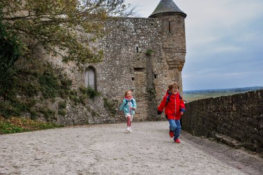 İki çocuk sevinçle eski bir şatonun yakınındaki tarihi bir taş patika boyunca koşuyor, manzaralı bir manzarayla çevrili.