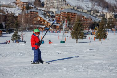 Karlı bir yamaçta, canlı kırmızı paltolu ve yeşil kasklı bir çocuk dağ evi tarzı bir kulübede kayak yapıyor. Tatillerde iyi eğlenceler.