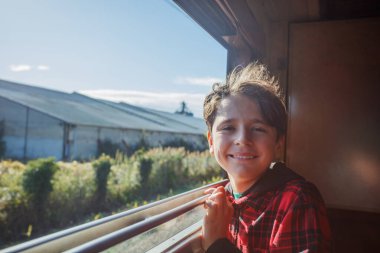 Çocuk kameraya gülümseyen pervazı kavrıyor, saçı rüzgarla savruluyor ve manzara arkasından süzülüyor.