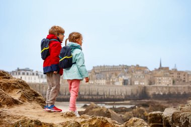 Sırt çantalı iki çocuk turist kayalık arazide dikilip Saint-Malo, Bretagne, Fransa 'nın uzak kasabasına bakıyorlar.