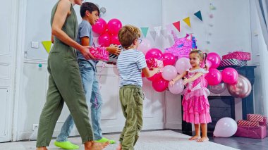 Şenlik sahnesinde pembe kostümlü bir kız iki erkek ve bir dizi renkli balonla annesinden hediye alıyor.