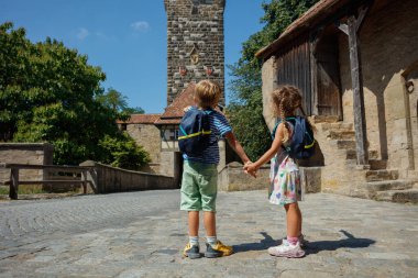 Sırt çantalı iki çocuk, eski bir taş binanın önünde el ele tutuşuyorlar.