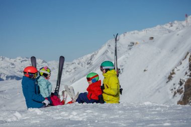 Bir grup kayakçı, anne, baba renkli miğferler ve ceketler giyen iki çocuk karlı bir dağın zirvesinde oturur ve karla kaplı manzarayı seyrederler.
