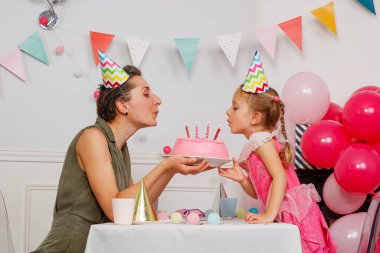 Kadın bir çocuğa mum ışığında pembe bir pasta sunuyor, birlikte mum üflüyorlar. İkisi de parti şapkası takıyor ve bölge balonlarla dolu.