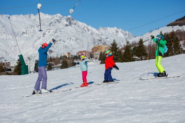 Bir kayak hocasının rehberliğinde, parlak kostümlü üç genç, beyaz, karlı bir tepede kayak yapıyor.