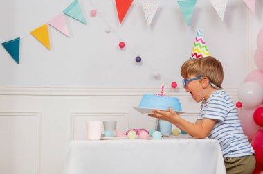 Gözlüklü ve parti şapkalı bir çocuk elinde renkli dekorasyonlarla çevrili mavi bir doğum günü pastasıyla bir şenlik masasında oturuyor.