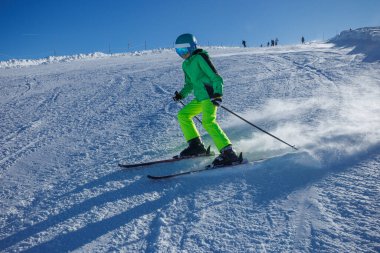 Canlı yeşil giyinmiş bir insan, karlı bir tepenin üzerinde kayak yapar ve açık bir arkaplanda son sürat döner.