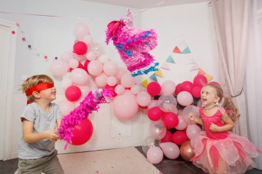 Bir parti sahnesinde, gözleri bağlı bir çocuk yanında neşeli bir kızla renkli bir pinataya yumruk atmak üzere.