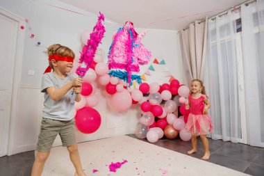 Bir çocuk, gözleri bağlı, tek boynuzlu atı yakalamaya çalışan bir yarasayı kavrıyor. Etrafı süslerle çevrili pembe bir piñata.