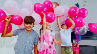 Üç çocuk, canlı pembe ve soluk renkli balonlarla çevrili bir partinin keyfini çıkarıyorlar.