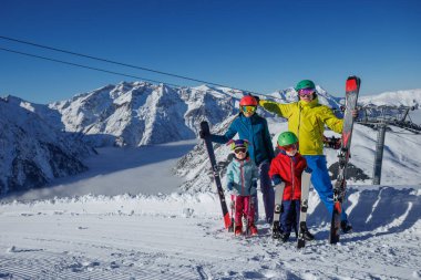 Dört anne, baba ve iki çocuklu bir aile, karlı bir dağ yamacında kayak malzemeleriyle poz veriyor, görkemli zirveler ve arka planda açık mavi bir gökyüzü var.