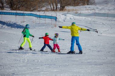 Kar yamacında kayak yapmayı öğrenen bir kayak hocası üç çocuğa rehberlik ediyor. Her biri parlak, renkli kış giysileri giyiyor.