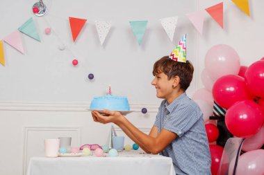 Genç bir çocuk, arka planda canlı bir masa balonunda otururken üzerinde mum olan turkuaz bir pastayı gösterirken sırıtıyor.