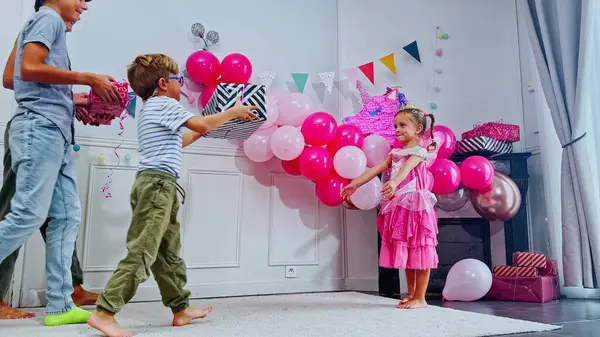 Neşeli bir kutlamada pembe elbiseli küçük bir kız iki erkekten bir hediye alır, anne, oda canlı balonlar ve çeşitli parti süsleriyle süslenir.