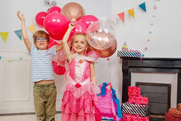Mavi gözlüklü, çizgili tişört giyen, pembe giyinmiş bir kızın yanında duran, neşeli balonlarla ve şenlikli dekorlarla çevrili, gülümseyen bir çocuk.