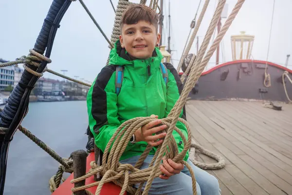 Canlı yeşil bir ceket giymiş genç bir çocuk ahşap bir gemi güvertesinde oturmuş, ipleri tutarken gülümsüyor.