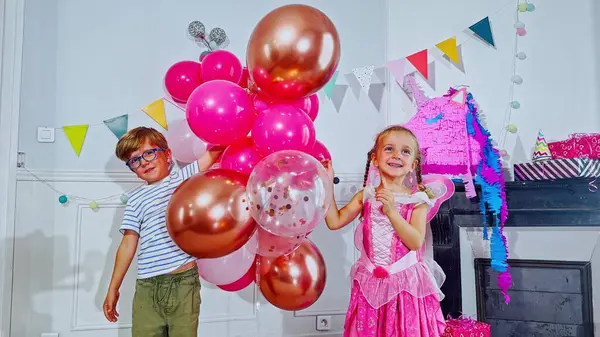 Genç bir erkek ve kız canlı pembe balonlarla dolu bir parti sahnesinde eğlenmek için çelenkleri kaldırırlar.