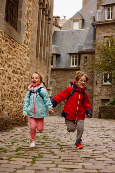 Parlak ceketler giyen iki çocuk, tarihi bir bölgede Saint-Malo, Bretagne, Fransa 'da taş binalar bulunan bir kaldırım taşından caddede neşeyle koşuyorlar.