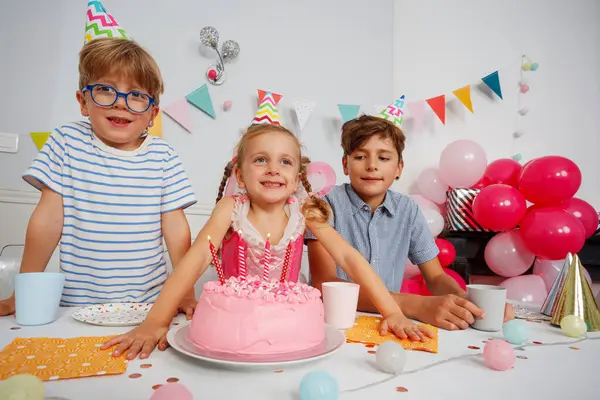 Neşeli bir partideki gençler, ayrılık şapkalarıyla süslenmiş ve canlı dekorlarla çevrili, pembe bir doğum günü pastası üzerinde gülümsüyorlar.