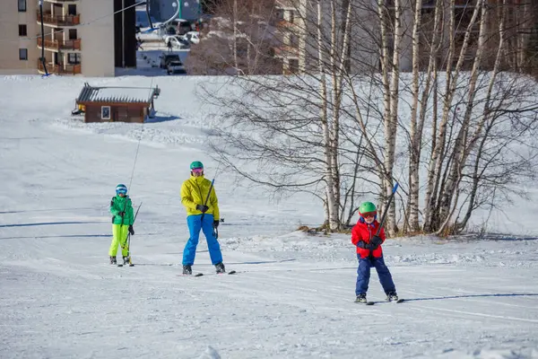 Çocuklar da dahil olmak üzere bir grup kayakçı, arka planda ağaçlar ve tatil köyleri bulunan karlı bir yamaçta kayak asansörü kullanıyor.