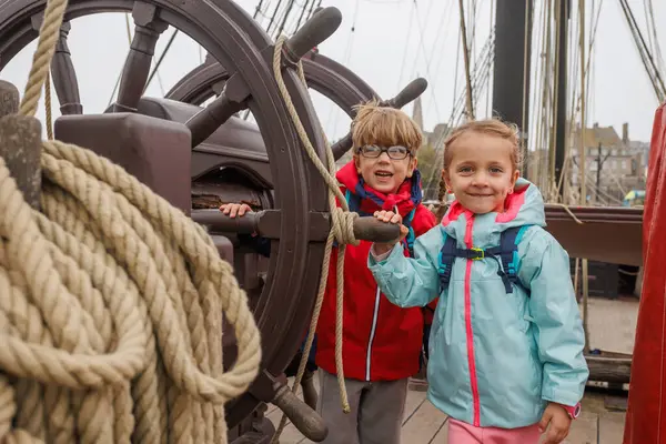 Ahşap bir güvertede, iki çocuk denizcilik iplerinin arasında, parlak dış giysiler giyip sırt çantaları taşıyorlar.