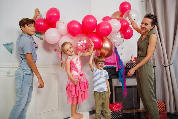 Süslü bir yerde pembe elbiseli bir kız tabureye çıkıp balon asıyor. Ailesi gülümseme dekorasyon odasının etrafında toplandı.