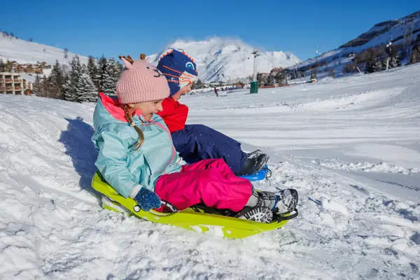 Kış giysileri içindeki hevesli çocuklar güneşli arka planda manzaralı binalarla karlı bir tepeden aşağı kaydılar.