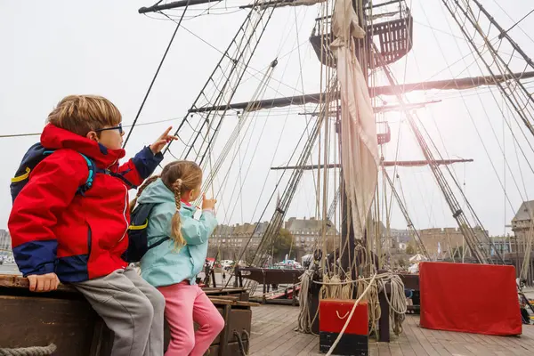 Kırmızı paltolu bir çocuk ve mavi ceketli bir kız büyük bir yelkenli gemisinin ahşap kutusunda, maceraya hazır bir şekilde oturuyorlar.