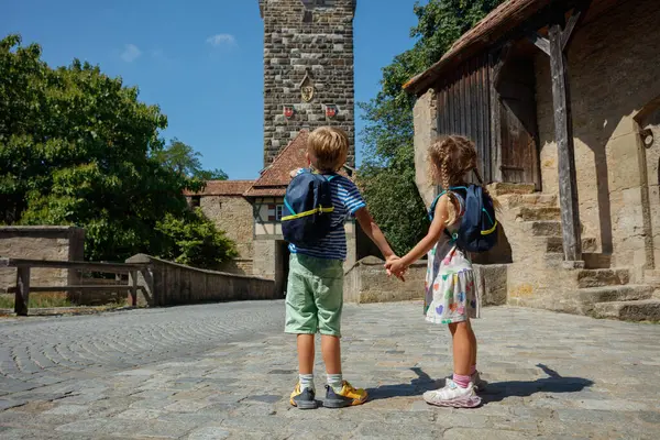 Sırt çantalı iki çocuk, eski bir taş binanın önünde el ele tutuşuyorlar.