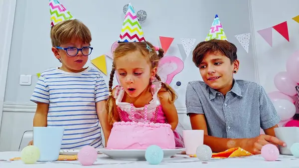 Bir doğum günü toplantısında, pembe elbiseli bir çocuk mumları üflüyor, iki arkadaşı sevinçten alkışlıyor.