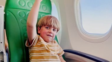 Uçakta, küçük bir çocuk mutlu bir şekilde oyuncak uçağıyla oynuyor, pencerenin yanındaki yeşil desenli koltuğun arka planında oturuyor.