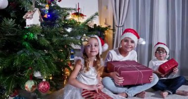 Konforlu oturma odasında, Noel Baba şapkalı üç genç güzel süslenmiş bir Noel ağacının yanında oturuyor, her biri hediyelerle dolu.