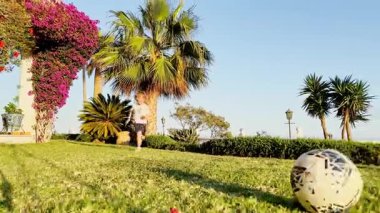 Küçük bir çocuk çimlere doğru koşuyor, bir futbol topuna doğru, etrafı palmiye ağaçlarıyla çevrili güneşli çimlerle çevrili